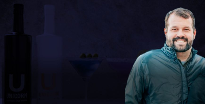Rick Hewitt, the founder of Unicorn Distillery, smiling in a jacket with two Unicorn Distillery bottles and cocktails in the background.
