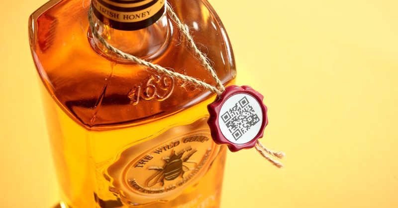 A close-up image of a bottle of "The Wild Geese Irish Honey" liqueur, featuring a QR code attached to the neck by a string with a faux wax red seal produced by Signet. The bottle is filled with a golden honey-coloured liquid, and the background is a warm yellow.