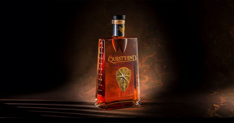 A dramatic image of a bottle of "Quest's End Paladin" bourbon whiskey. The bottle has a unique shape and is adorned with a shield emblem and a neck wrap, both produced by Signet. The background is dark with a hint of golden light, highlighting the rich amber colour of the whiskey.