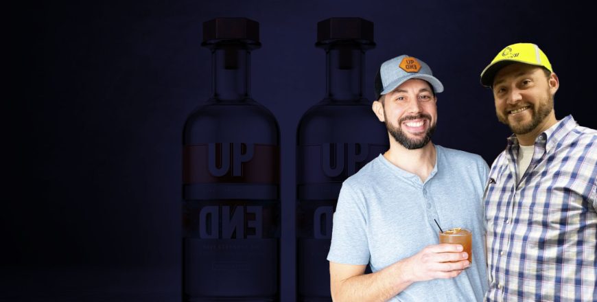 Matthew McKee & Chris Lofrese posing with a drink in front of UP END Navy Strength Gin bottles, wearing branded hats and casual shirts, promoting UpEnd Distilling