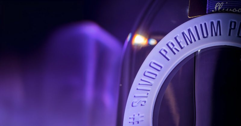 The image shows a close-up view of the top section of a bottle with a prominent plastic ring around its neck. The ring has the text "SLIVOO PREMIUM PLUM DISTILLATE" embossed on it. The background is a gradient of purple and dark blue, creating an elegant and sophisticated look.